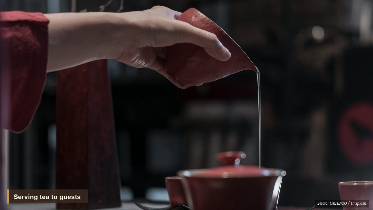 A hand pouring tea from a red gaiwan beside a plum blossom branch