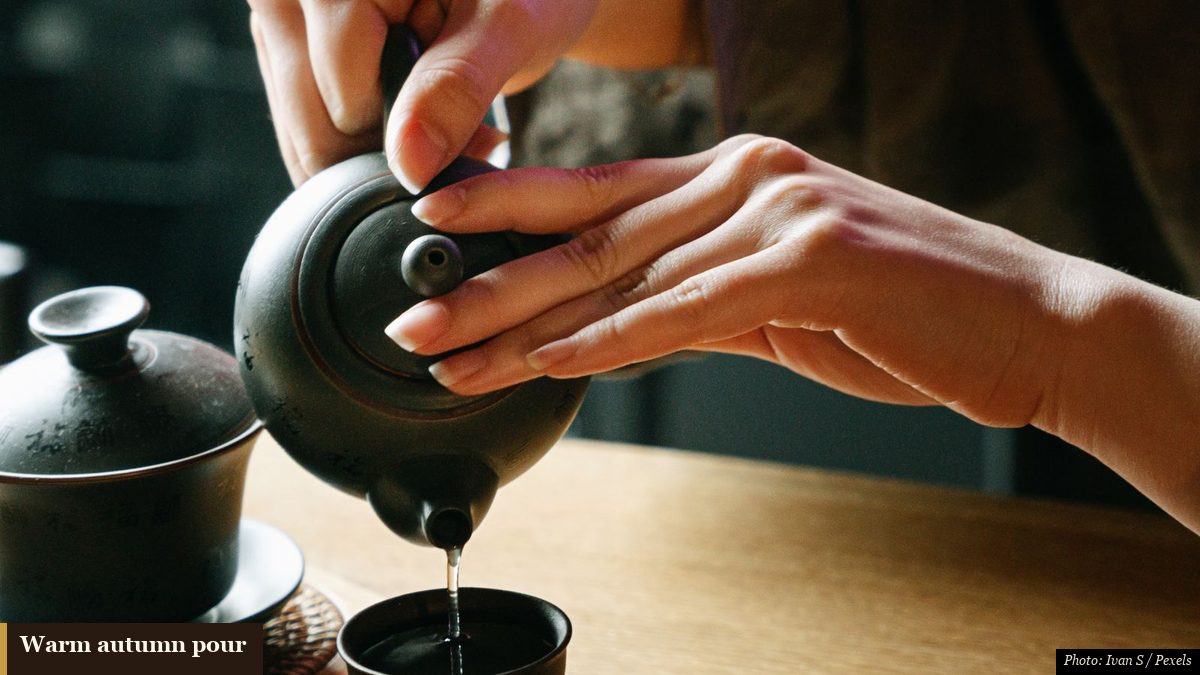 Hands pouring dark roasted tea into a small cup on a bamboo tea tray