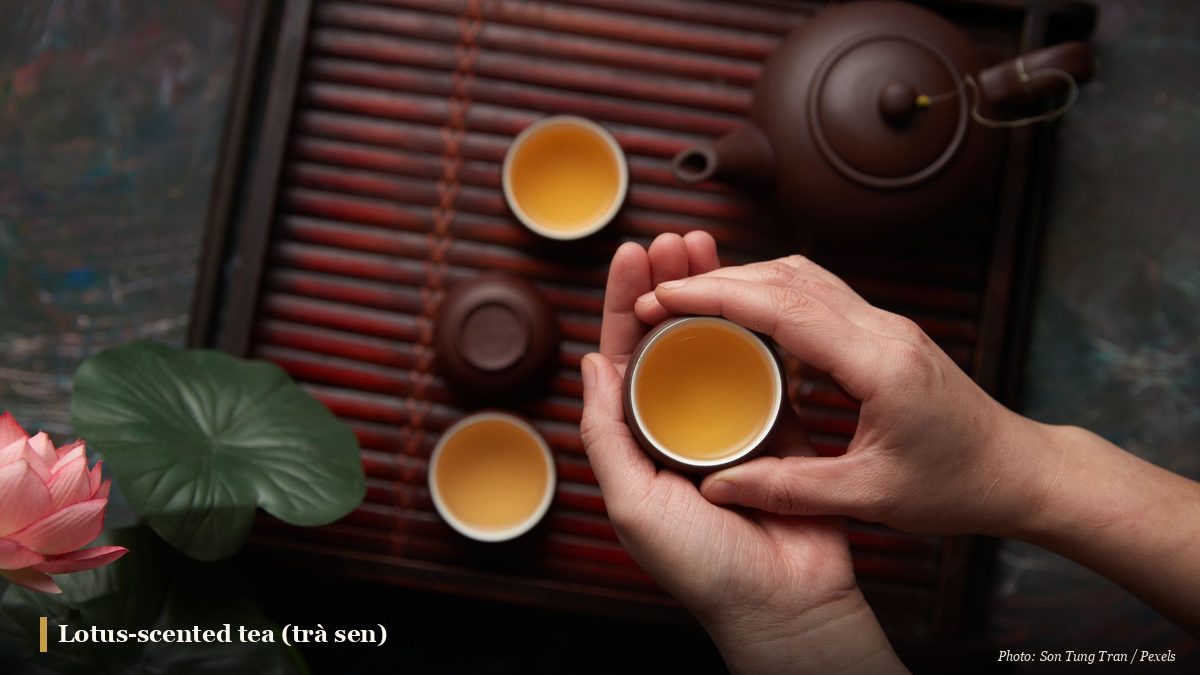 Hands holding a cup of golden lotus tea beside a clay teapot and a lotus flower