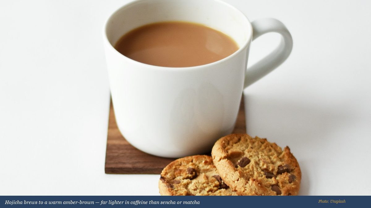 Warm amber hojicha brew in a rustic ceramic cup beside a white teapot on a wooden tray