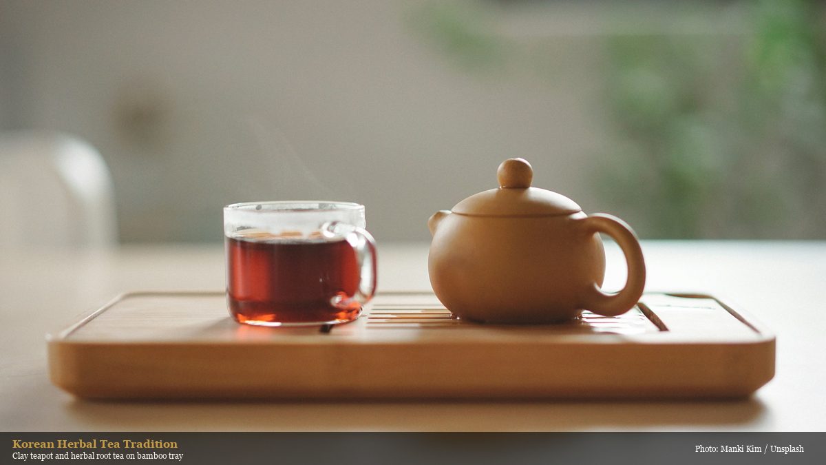 Korean herbal root tea in a glass cup alongside a clay teapot on a bamboo tray, showing the tradition of root and herb tea preparation