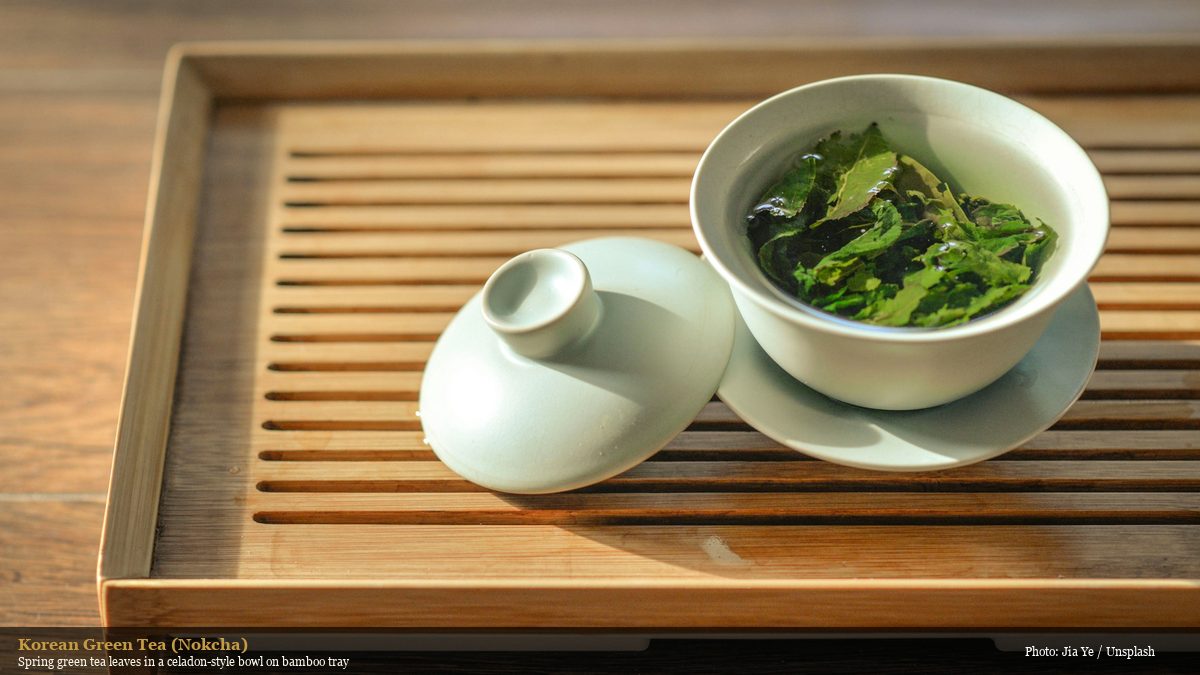Fresh green tea leaves in a white celadon-style bowl on a bamboo tea tray, representing Korean nokcha green tea tradition