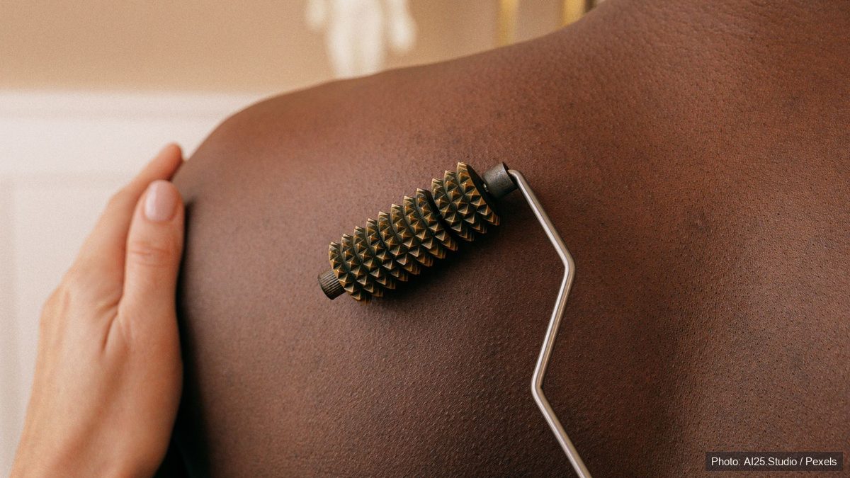 Therapist rolling a wooden massage tool along a client's back with an anatomical model nearby