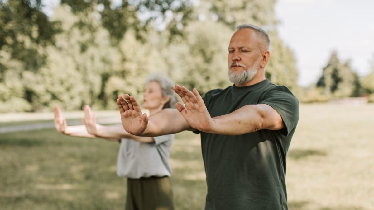 Older couple practicing qigong with palms extended forward in a green park