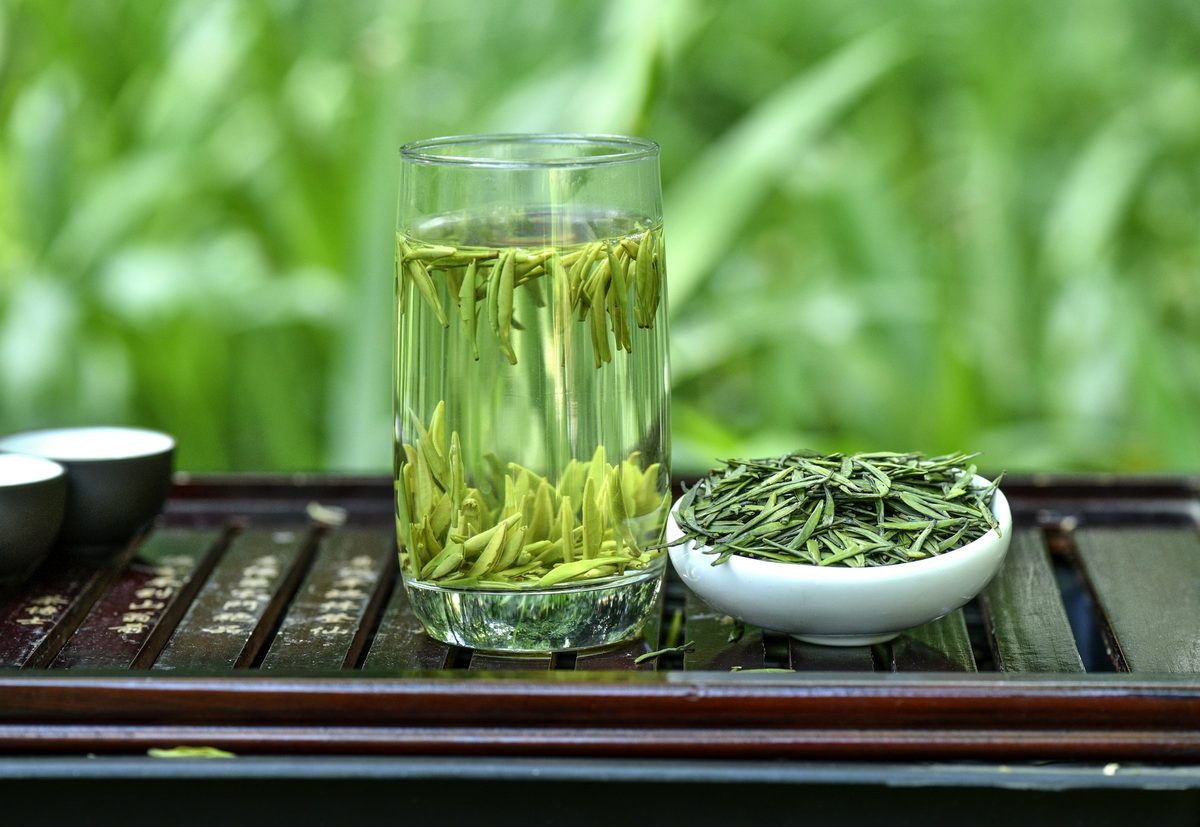 A clear glass cup of freshly brewed Japanese green tea with bright green leaves visible, photographed on a light background with fresh herbs