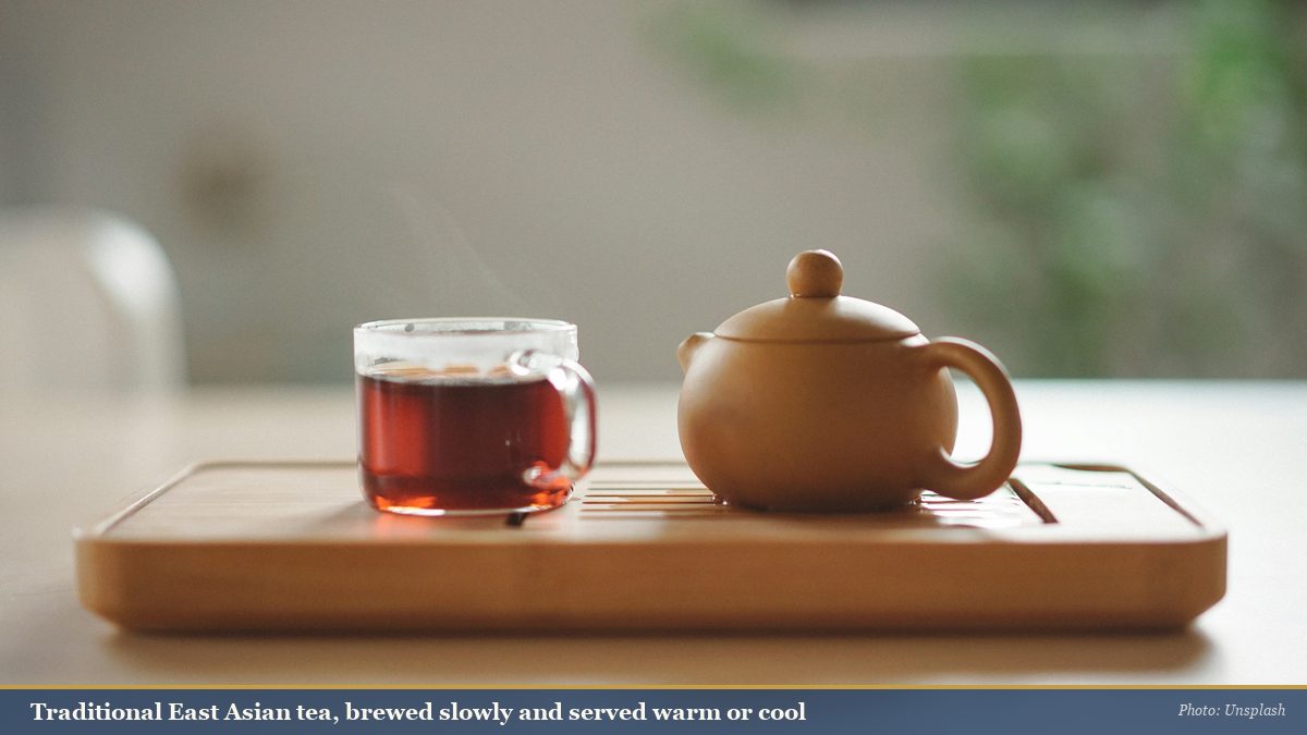 A glass cup of amber tea beside a clay teapot on a wooden tea tray