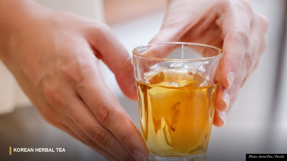 Hands holding a glass of golden Korean herbal citron tea beside bowls of dried botanicals