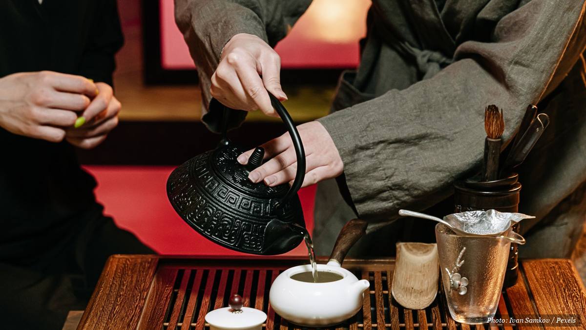Hands pouring hot water from an iron kettle into a small teapot during a tea ritual