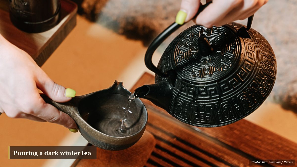Hands pouring dark tea from a cast-iron teapot into a small clay cup on a wooden tray