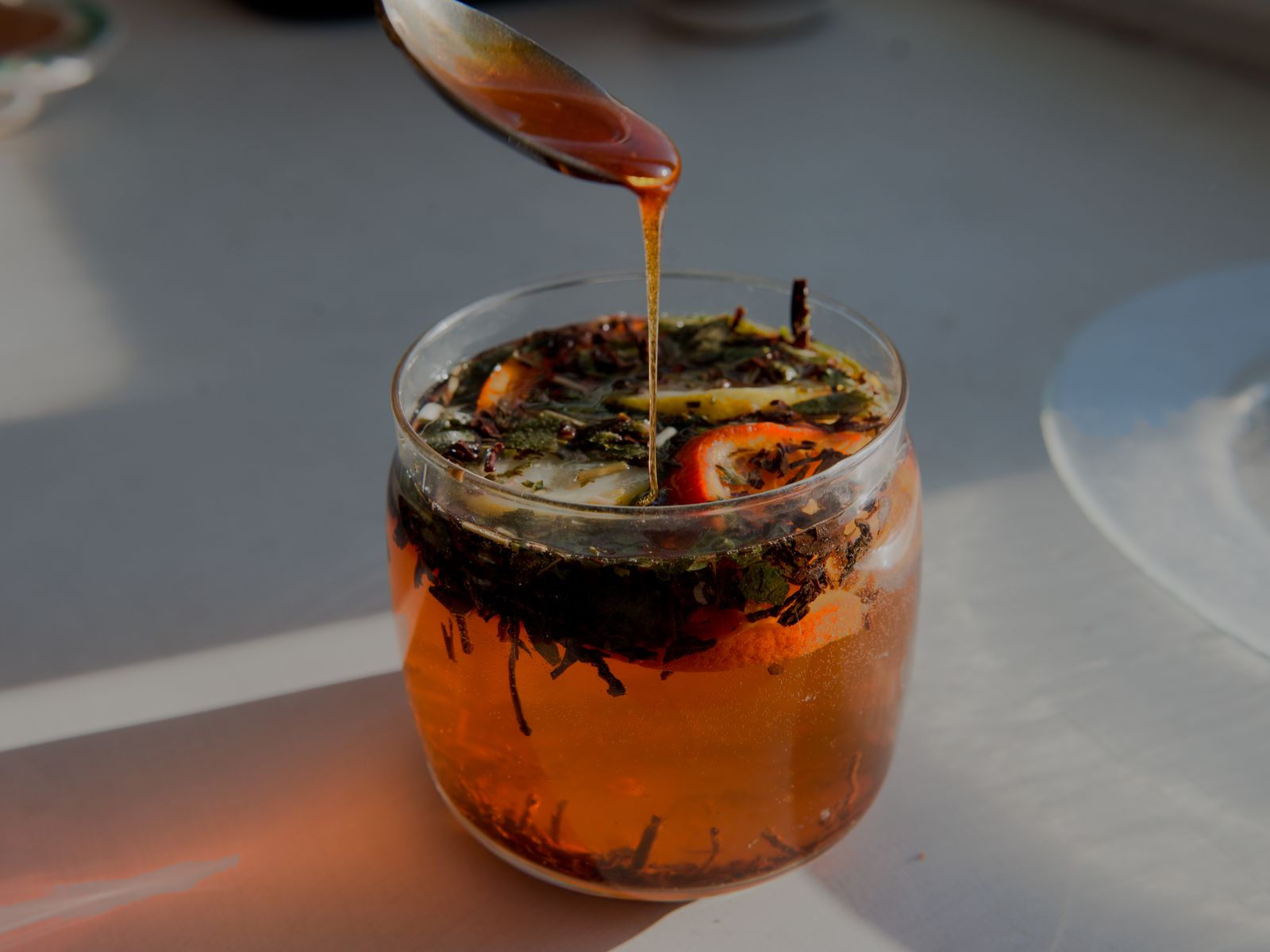 Honey being drizzled into a warm herbal citrus tea infusion in a glass jar, rich amber tones