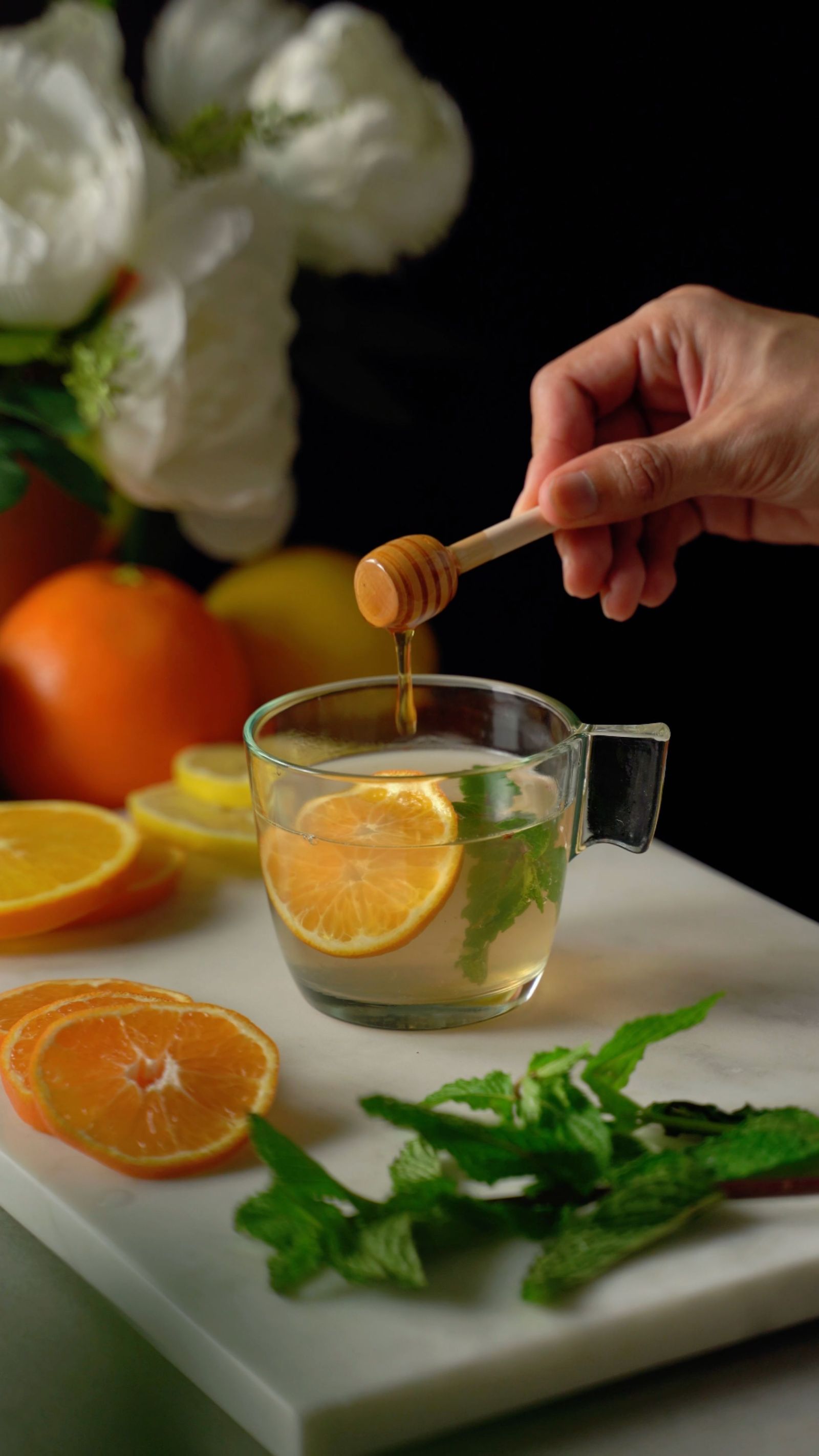 Hand holding honey dipper over a glass mug of warm citrus tea with orange slices and mint, surrounded by fresh citrus fruits